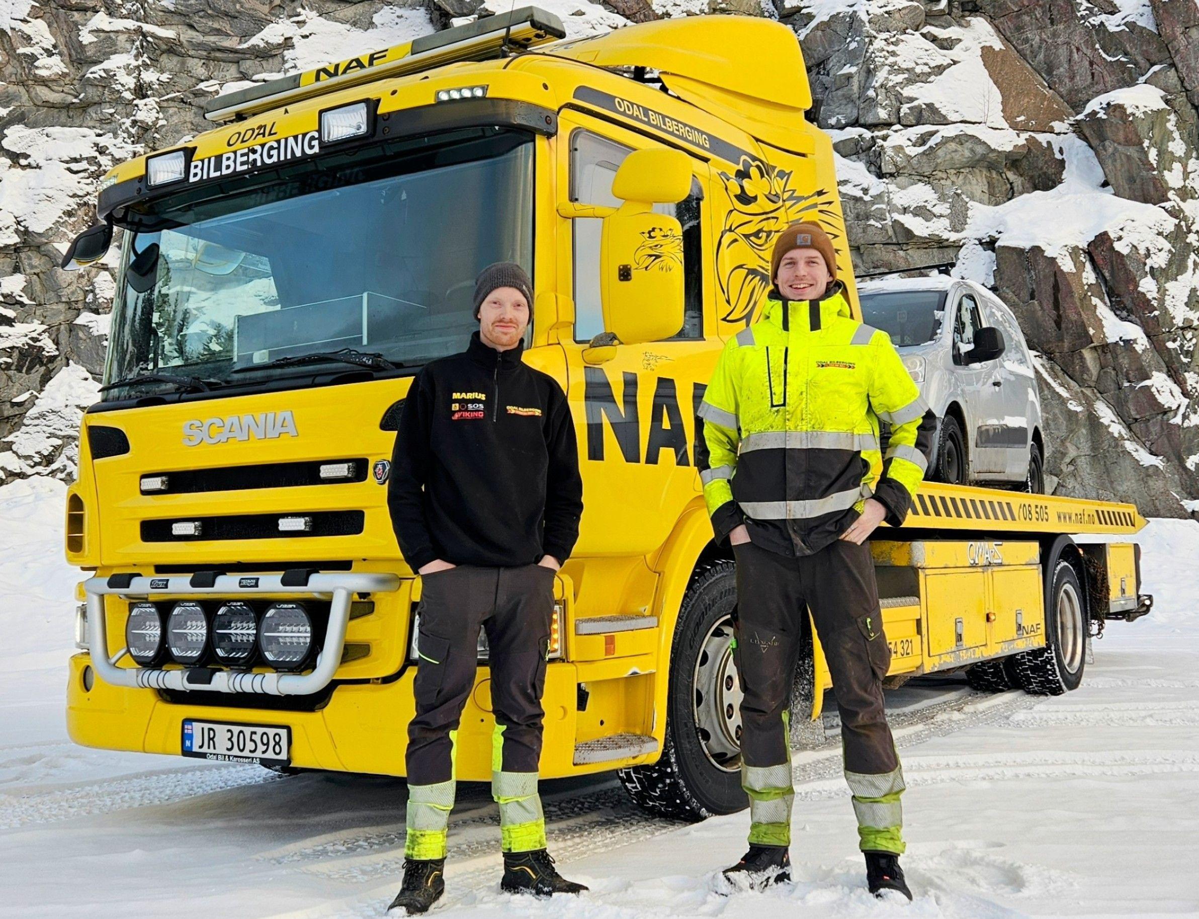 To menn står foran en gul bergingsbil med en bil på slep, omgitt av snø og steiner.
