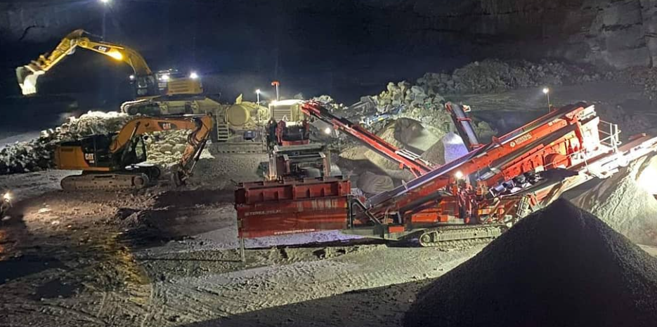 Gravemaskiner og transportbånd i en gruve, med stein og grus i nattemørket. Belysning fra maskinene skaper en dramatisk effekt.