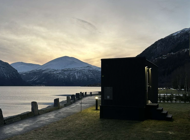 En moderne svart hytte ved vannet med snødekte fjell i bakgrunnen under en solnedgang.