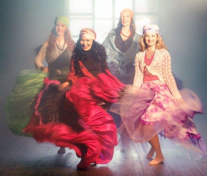 Fire kvinner i fargerike kjoler danser med lange skjørt i et svakt opplyst rom.