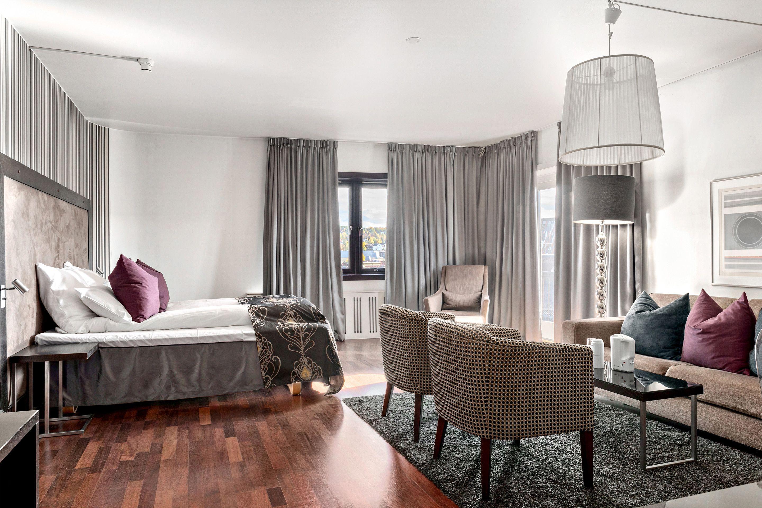Lyst og moderne hotellrom med dobbeltseng, stoler, sofa og store vinduer med gardiner.
