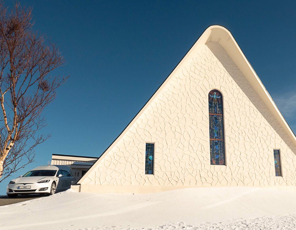 Moderne kirke med spiss takform, hvit fasade og fargerike vinduer, omgitt av snø og klar blå himmel.