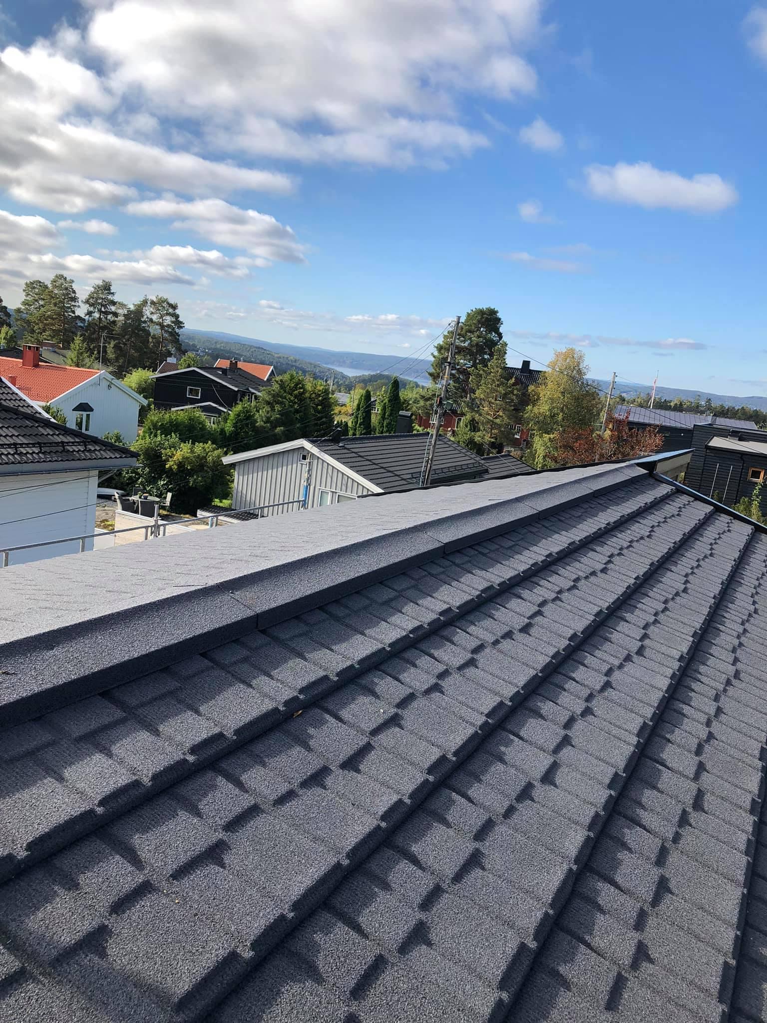 Utsikt fra taket med grå takplater, hus og trær i bakgrunnen under en blå himmel.