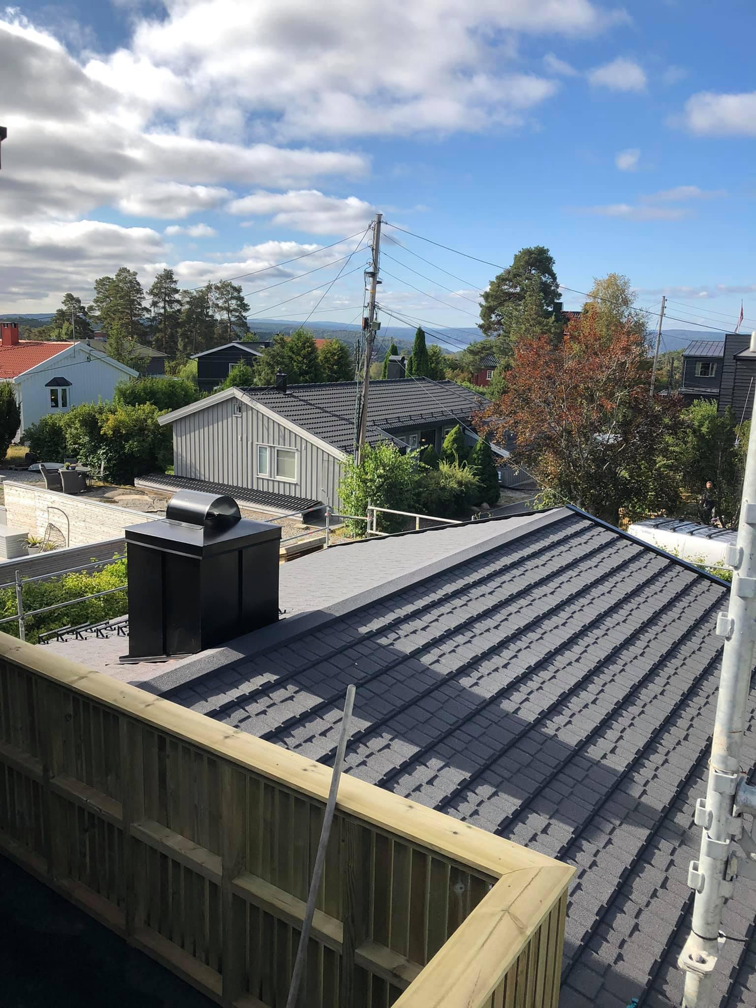 Utsikt over takene på hus i et nabolag med trær og blå himmel i bakgrunnen.