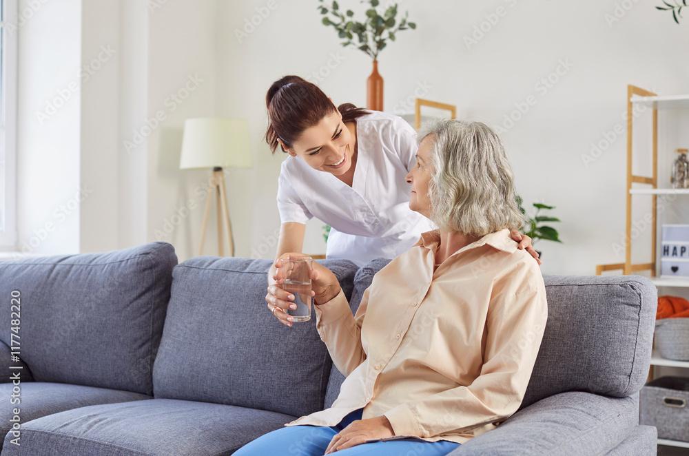 En omsorgsfull sykepleier snakker med en eldre kvinne på en sofa i et lyst rom.
