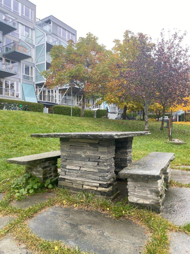 Et steinbord med benker i en park, omringet av trær med høstfarger og moderne bygninger i bakgrunnen.