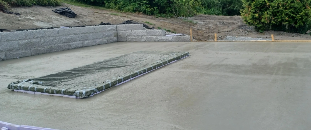 Betongfundament på byggeplass med en del av overflaten dekket av plast for tørking. Grønt område i bakgrunnen.