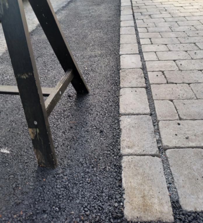 Asfaltert vei med brostein og et svart arbeidsstativ i forgrunnen.