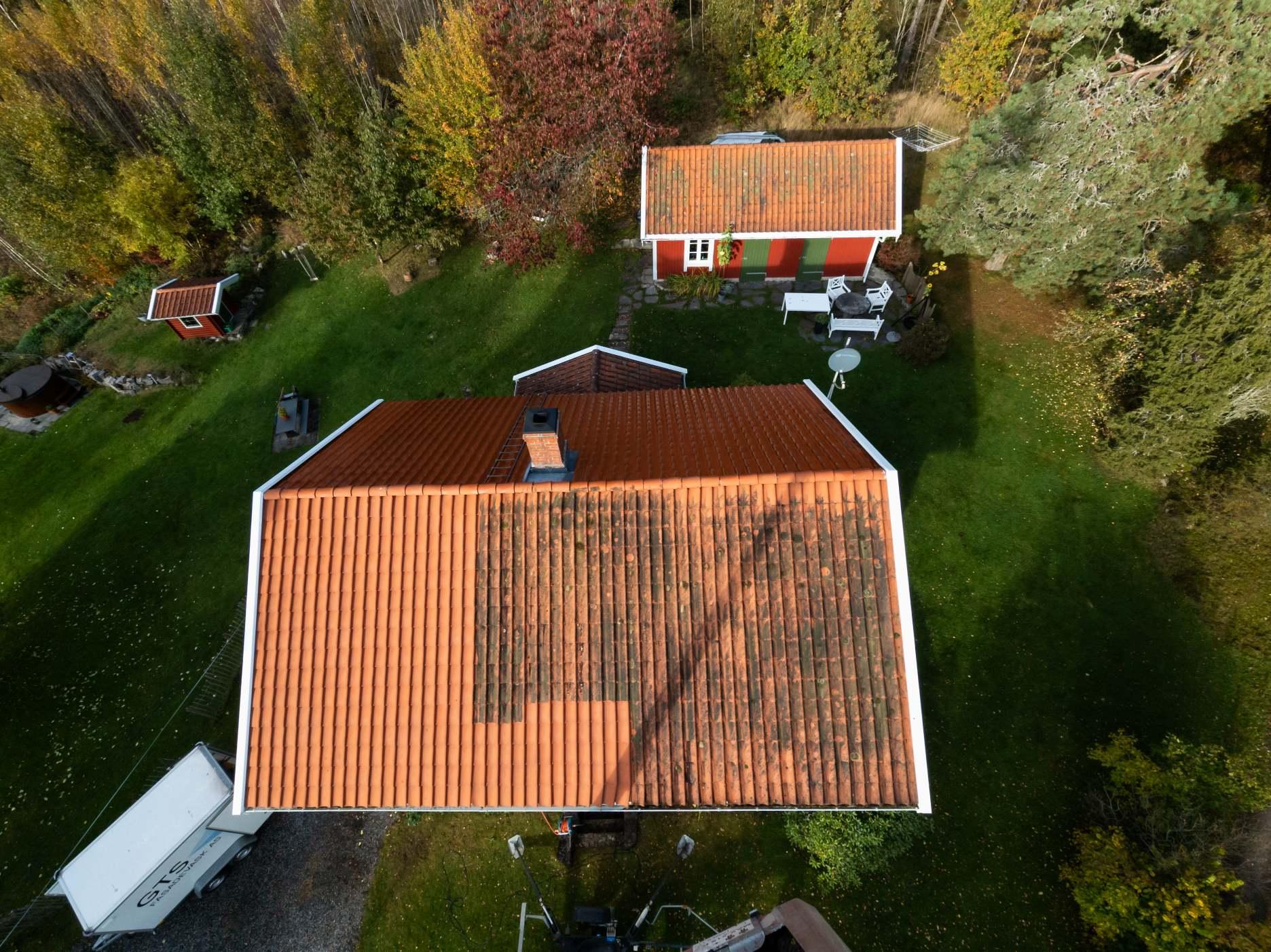 Oversikt over taket på et hus med rød hytte i bakgrunnen, omringet av grønt gress og trær.