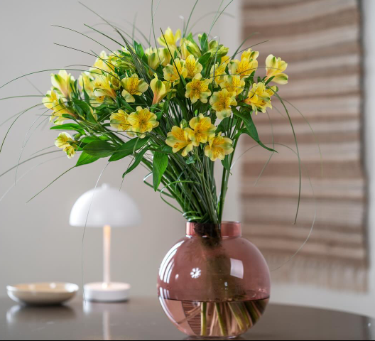 En bukett med gule alstromeria blomster i en rosa vase, plassert på et bord med en lampe i bakgrunnen.