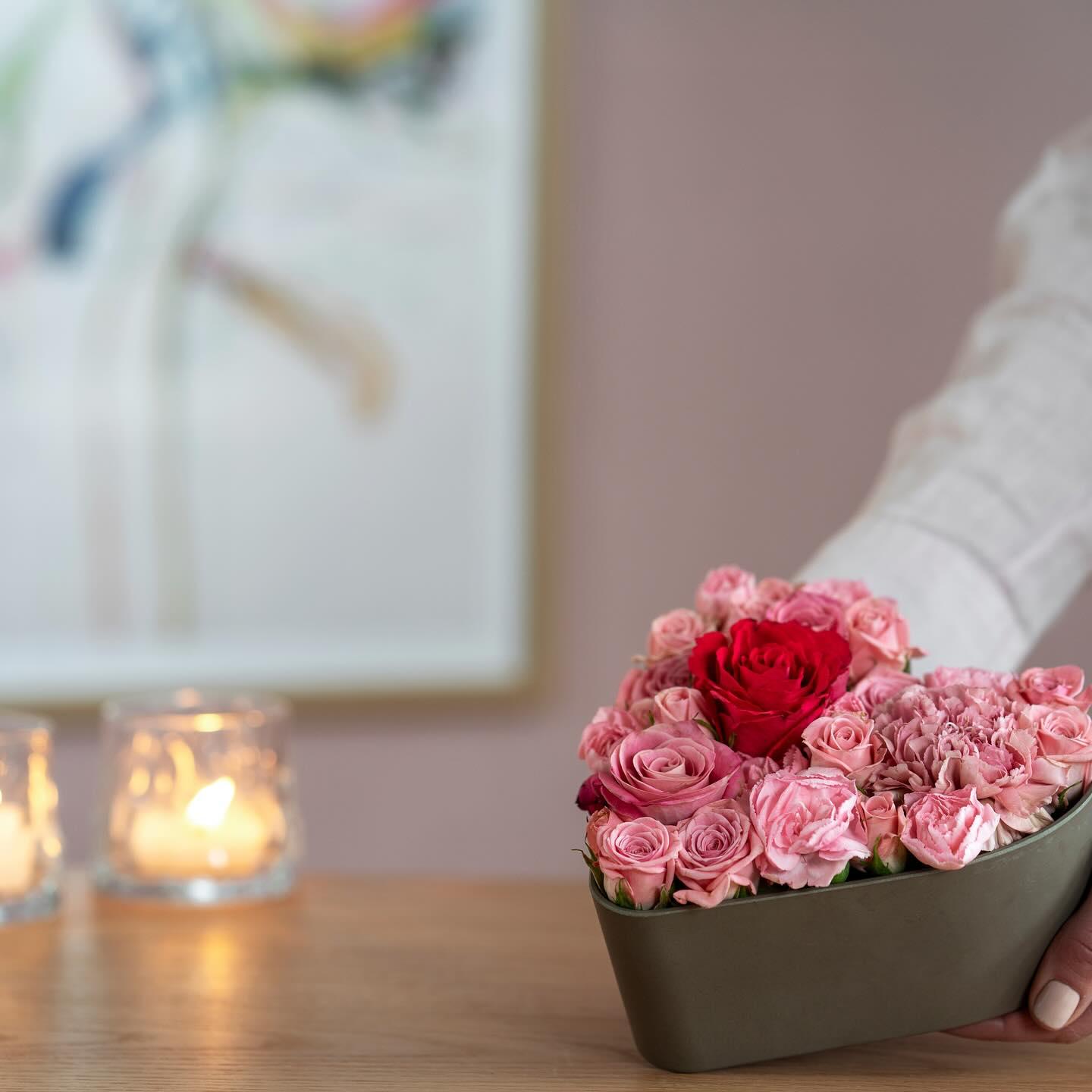 En hånd holder en hjerteformet blomsterdekorasjon med rosa roser og en rød rose, med stearinlys i bakgrunnen.