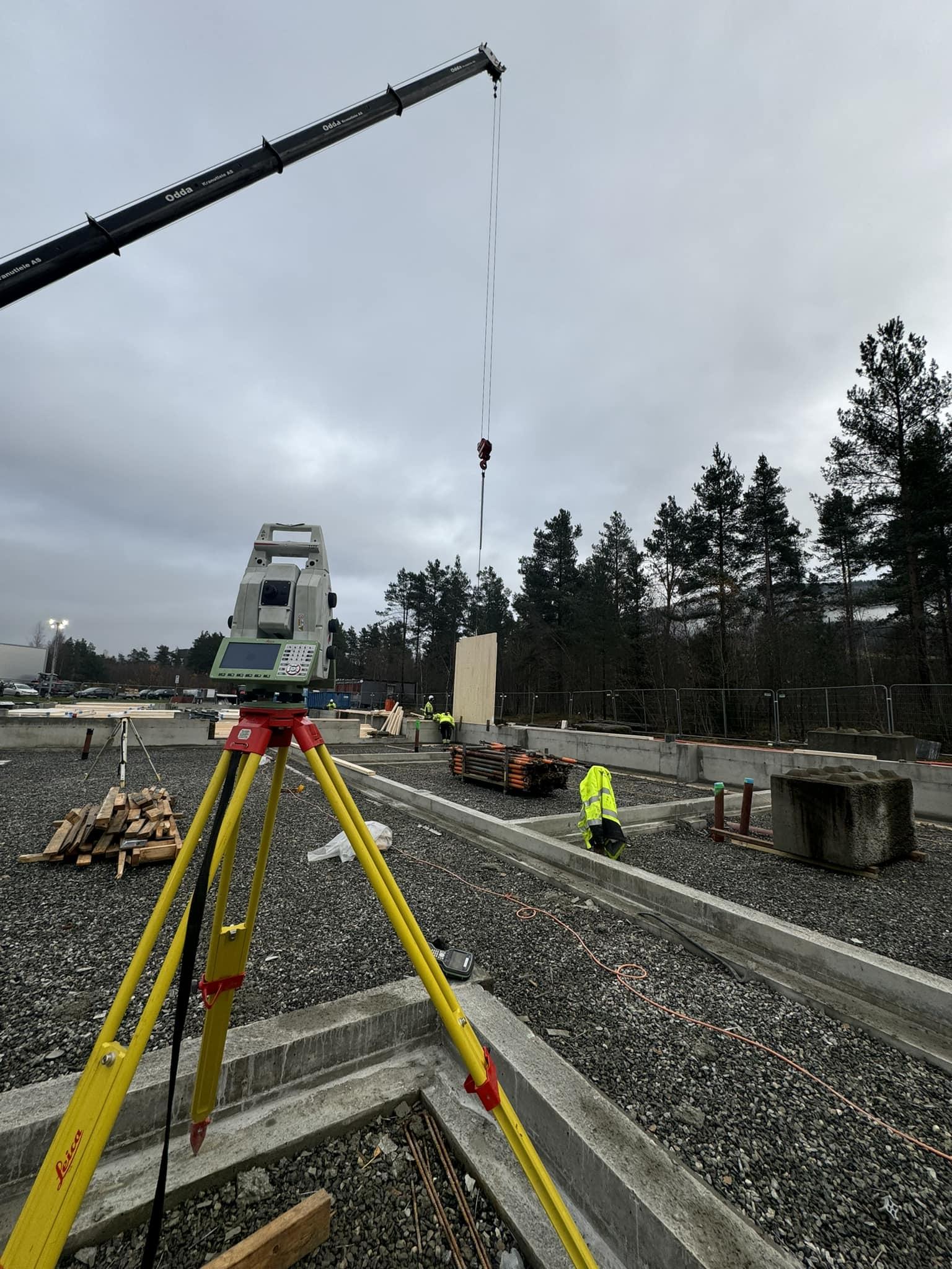 Kran løfter en stor betongvegg på byggeplass med måleutstyr i forgrunnen og trær i bakgrunnen.