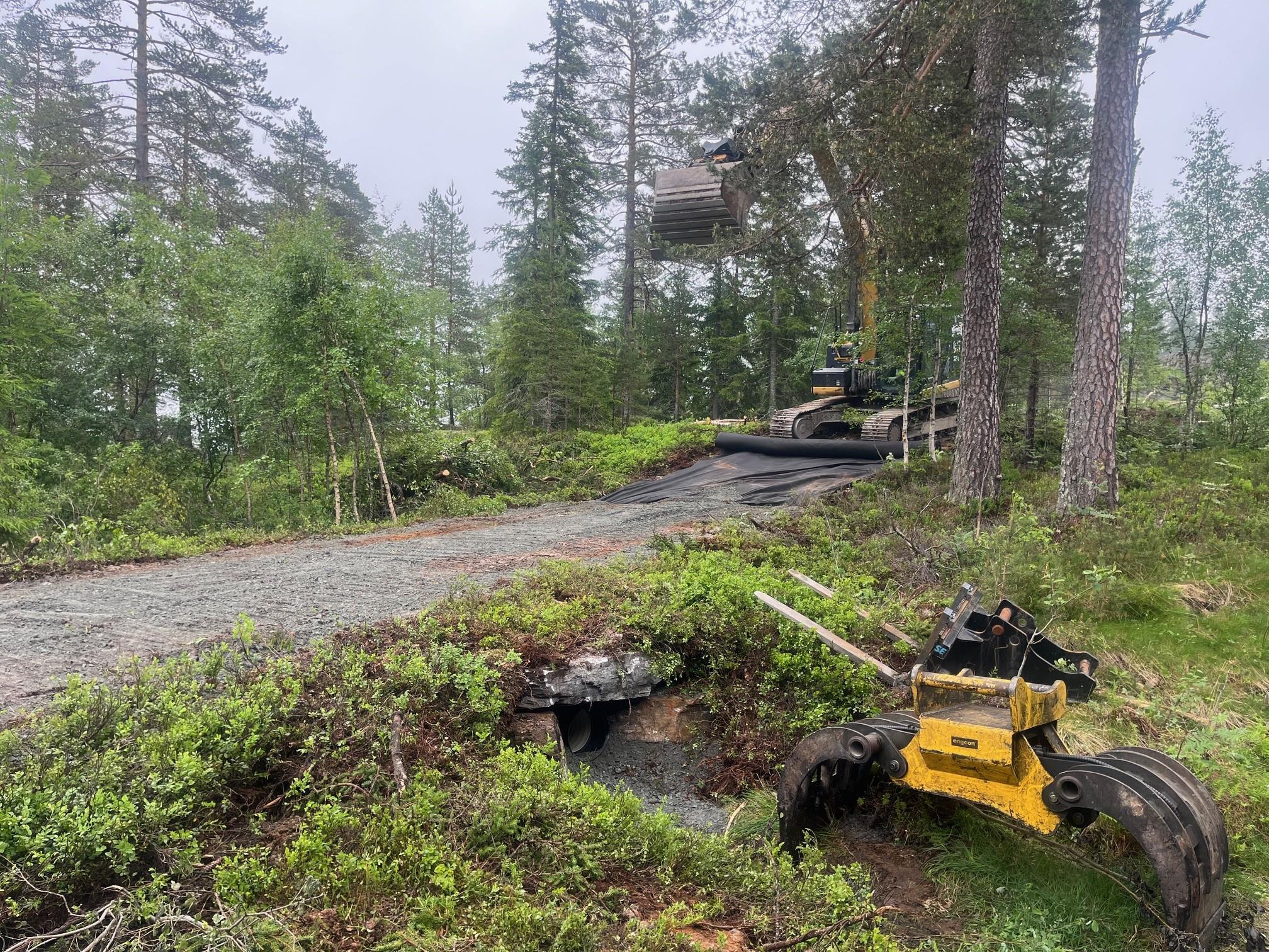 En grusvei med skog på begge sider, en gravemaskin i bakgrunnen og en gummihjulsmaskin i forgrunnen.