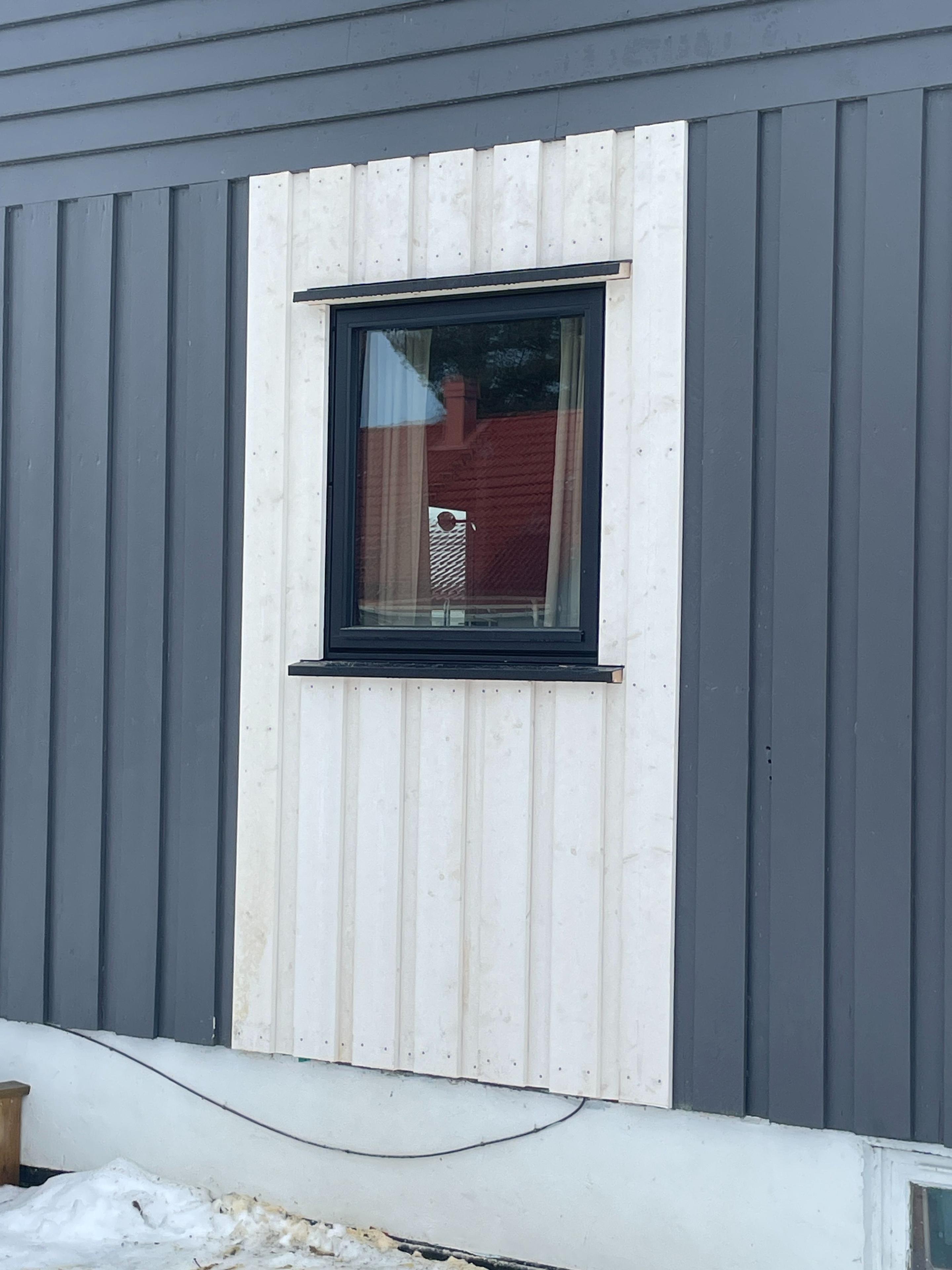 Vindu med svart ramme på en grå og hvit kledningsvegg, delvis dekket av snø.