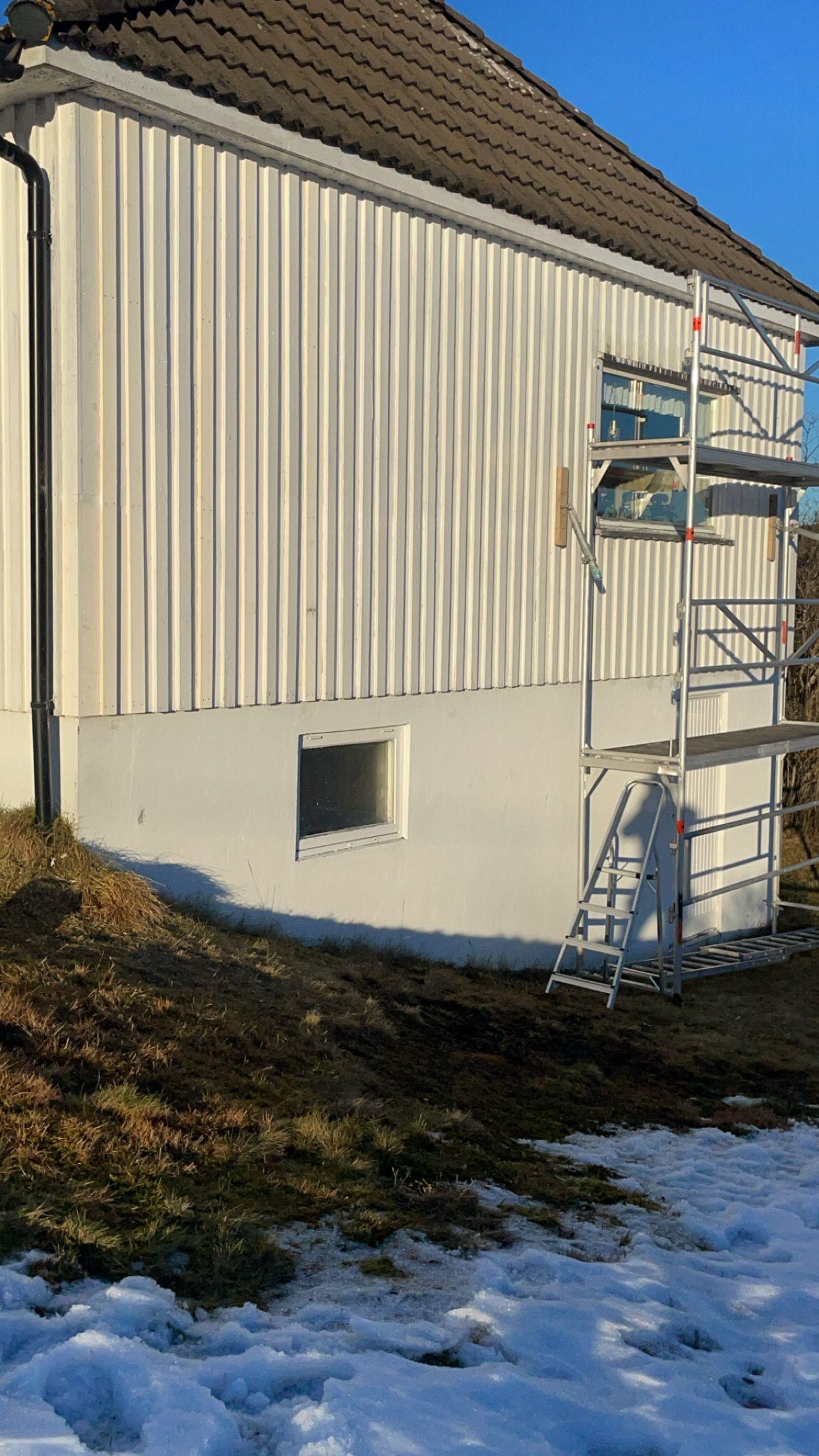 Hvit husvegg med stillas og snø i hagen, under solfylt himmel.