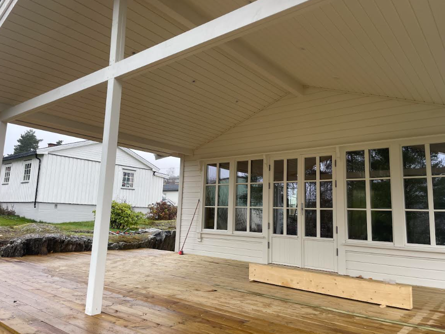 Moderne hytte med stor terrasse, hvite vegger og glassdører, omgitt av natur og andre bygninger.