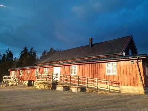 Rødt trehus med skråtak og veranda, omgitt av trær under en blå himmel.