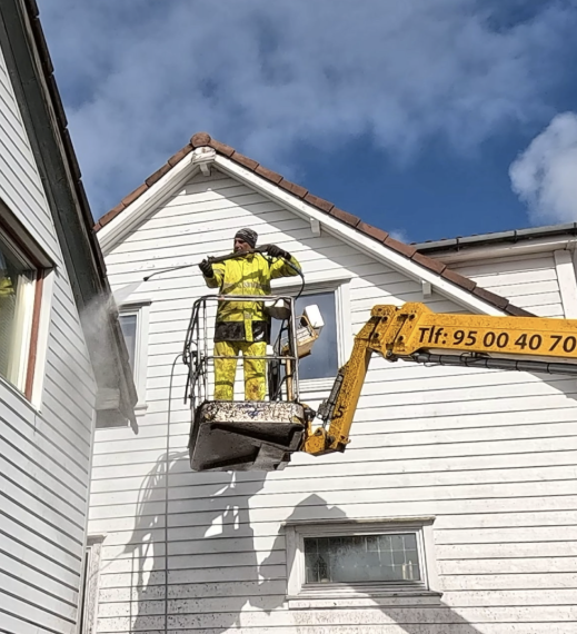 En arbeidstaker i verneutstyr bruker en lift for å vaske veggen på et hus. Blå himmel i bakgrunnen.