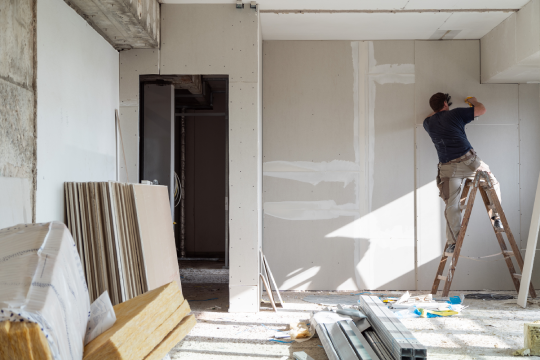 En håndverker som jobber med gipsplater på en stige i et rom under oppussing. Byggematerialer ligger på gulvet.