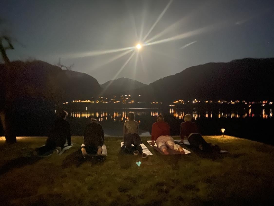 Fem personer sitter på en gressplen ved vannet om natten, med månen som lyser over fjellene.