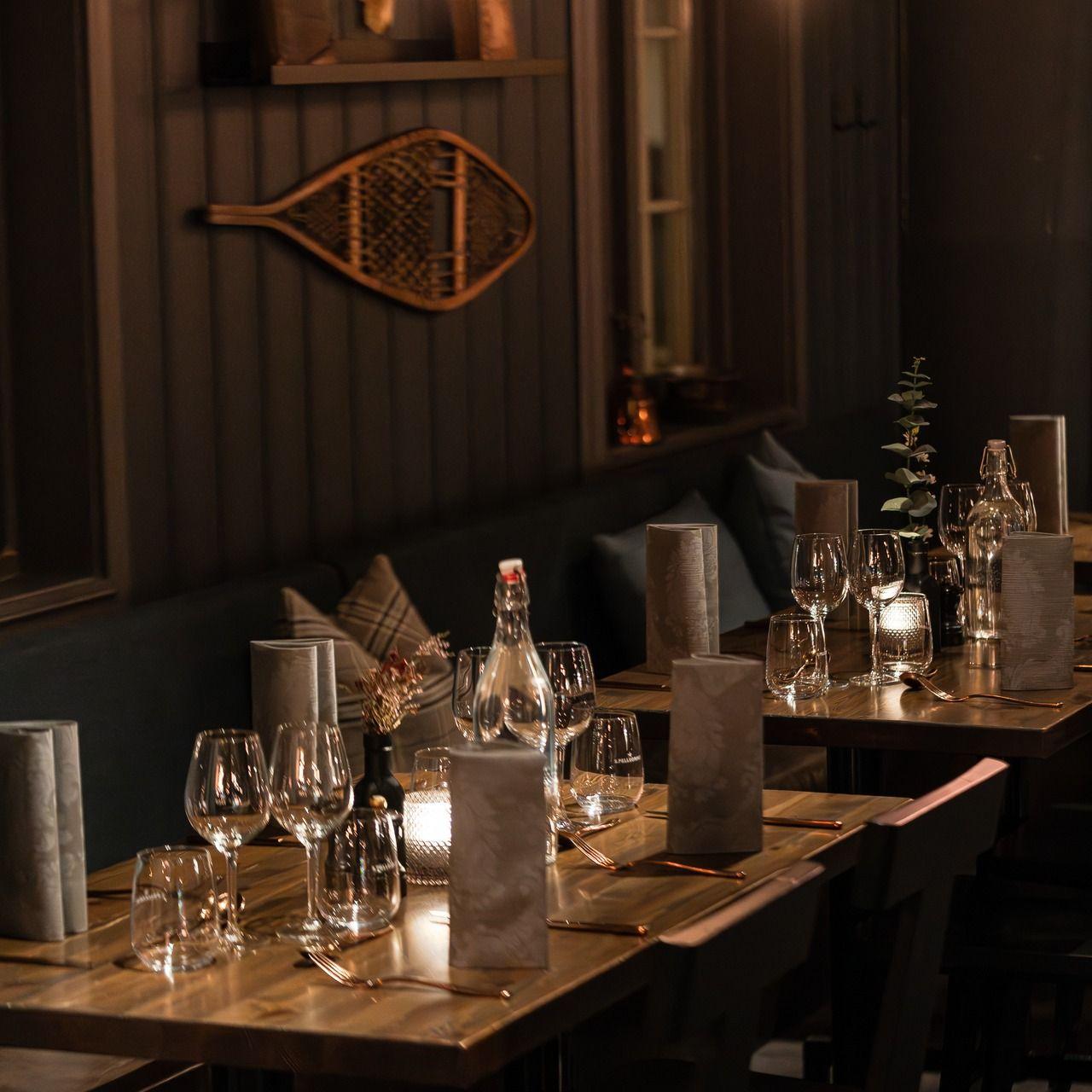 Koselig restaurantinteriør med bord dekket med glass og menyer, dempet belysning og dekorative elementer.
