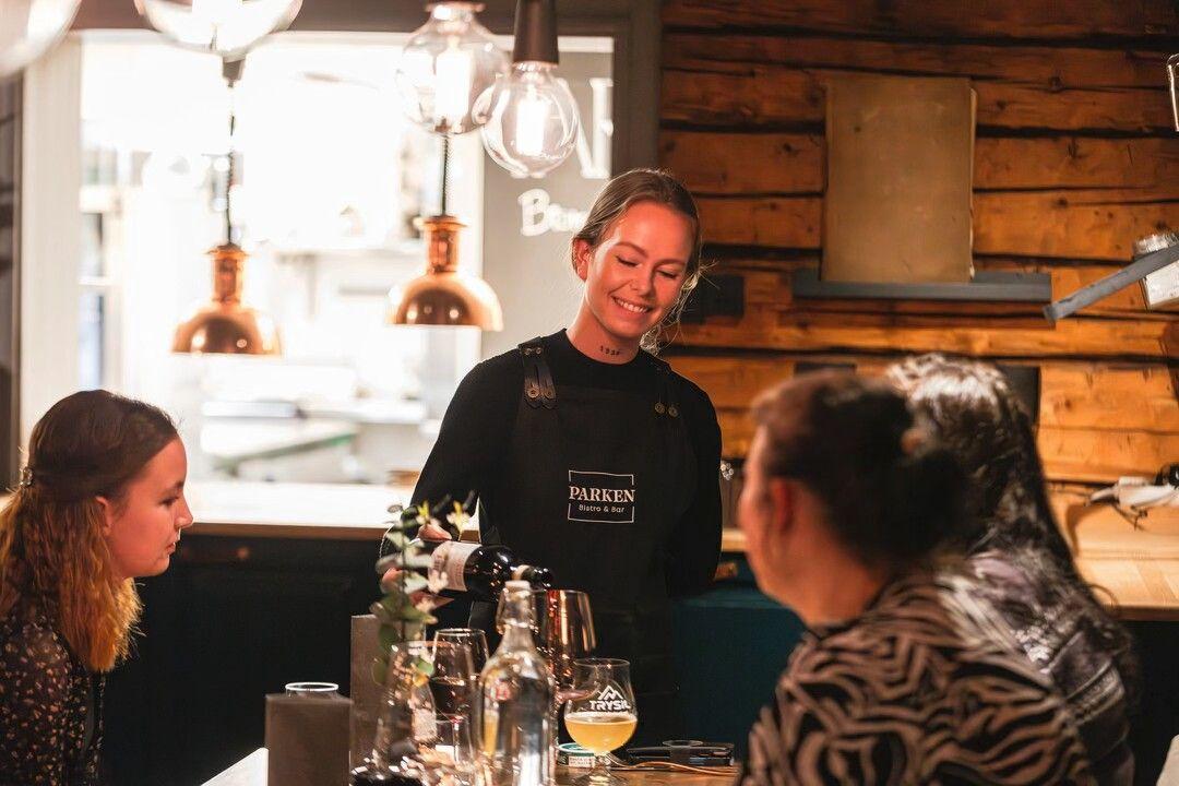 En smilende servitør serverer drinker til gjester i en koselig restaurant med trevegger.