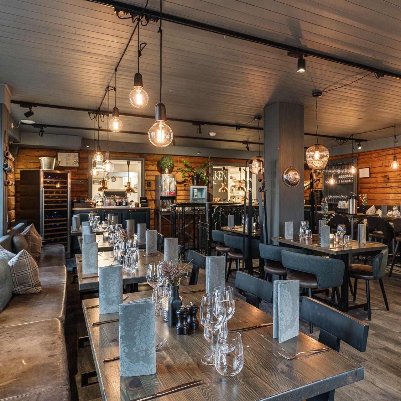 Moderne restaurantinteriør med trevegger, stilige lamper og bord dekket med glass og servise.
