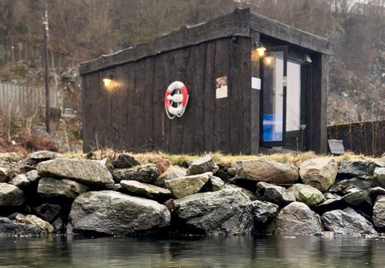 En liten, svart hytte ved vannet med en redningskrans på veggen og steiner rundt.