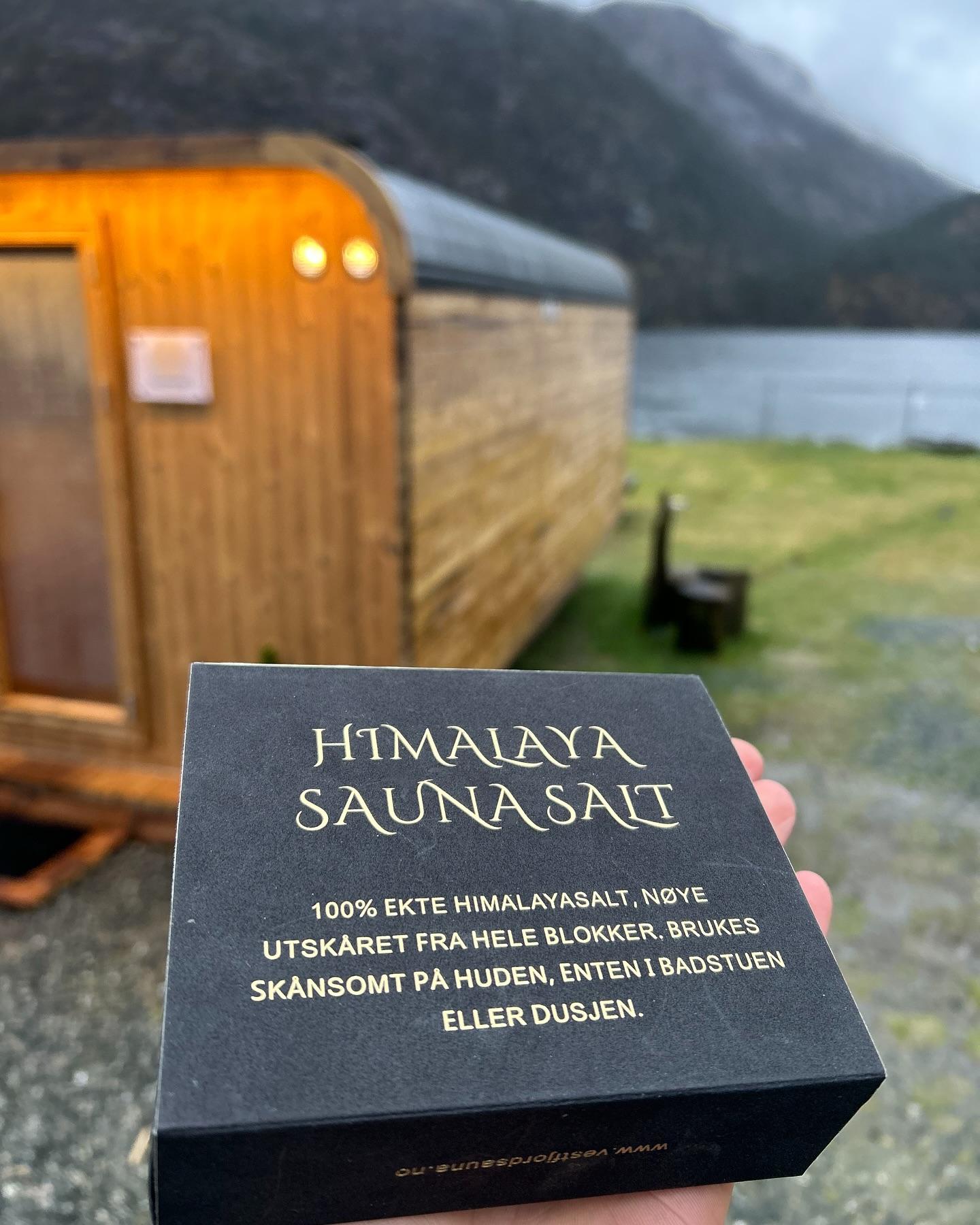 Boks med Himalaya saunasalt foran en liten hytte ved vannet, med fjell i bakgrunnen.