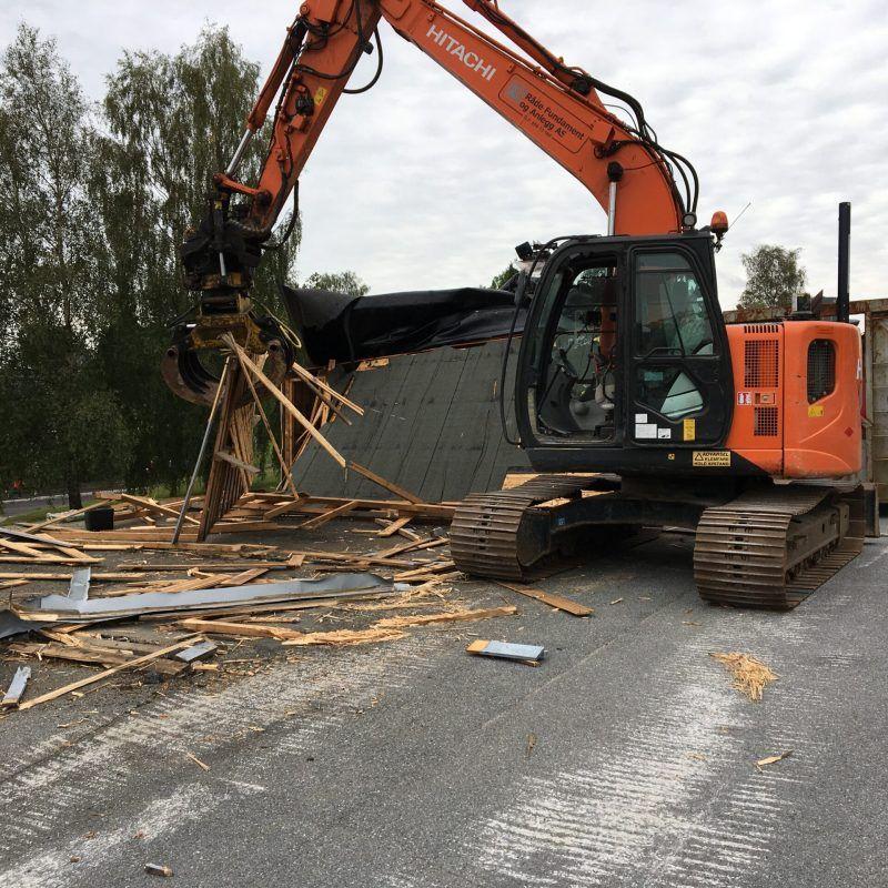 Gravemaskin fjerner bygningselementer fra en byggeplass med treverk og skrot spredt rundt.