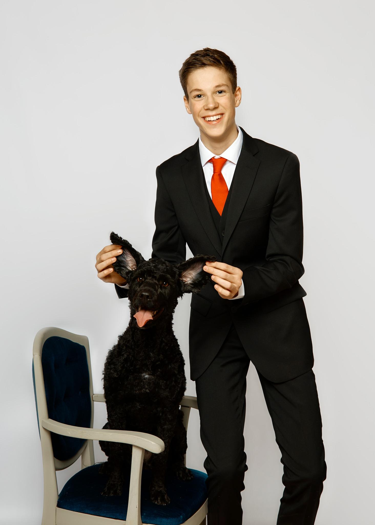 Ung mann i dress smiler mens han holder en svart hunds ører, sittende på en stol.