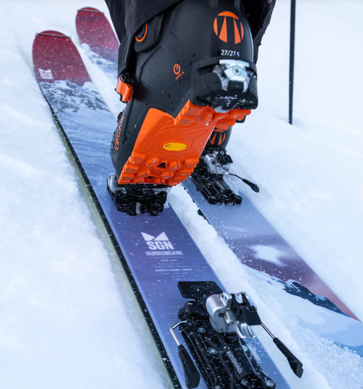 En skistøvel i oransje og svart på ski i snøen, med skiutstyr synlig i bakgrunnen.