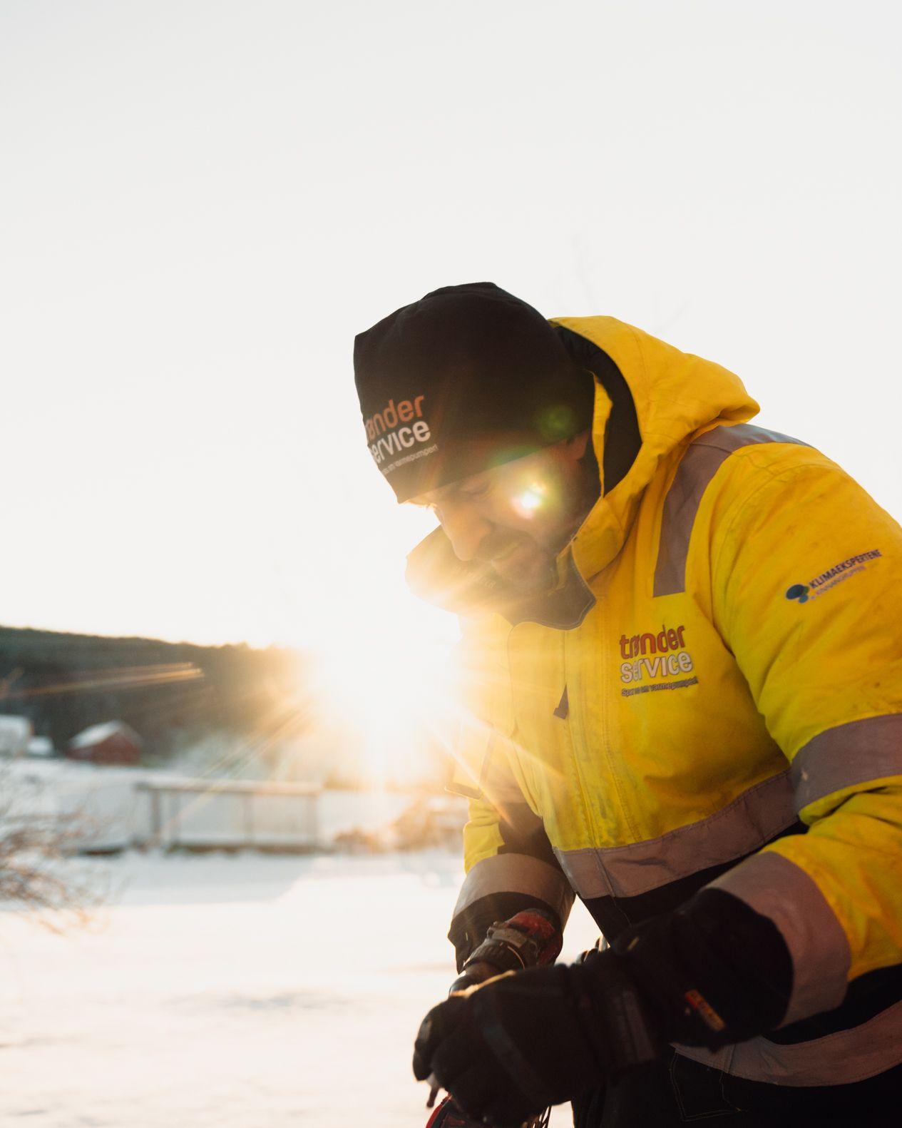 En arbeidstaker i gul vinterjakke og lue jobber ute i snøen med sollys i bakgrunnen.