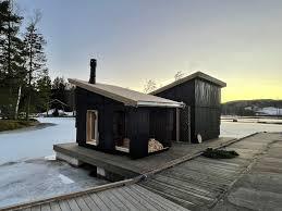 En svart hytte ved vannet med en tredekk og snødekte omgivelser i solnedgang.