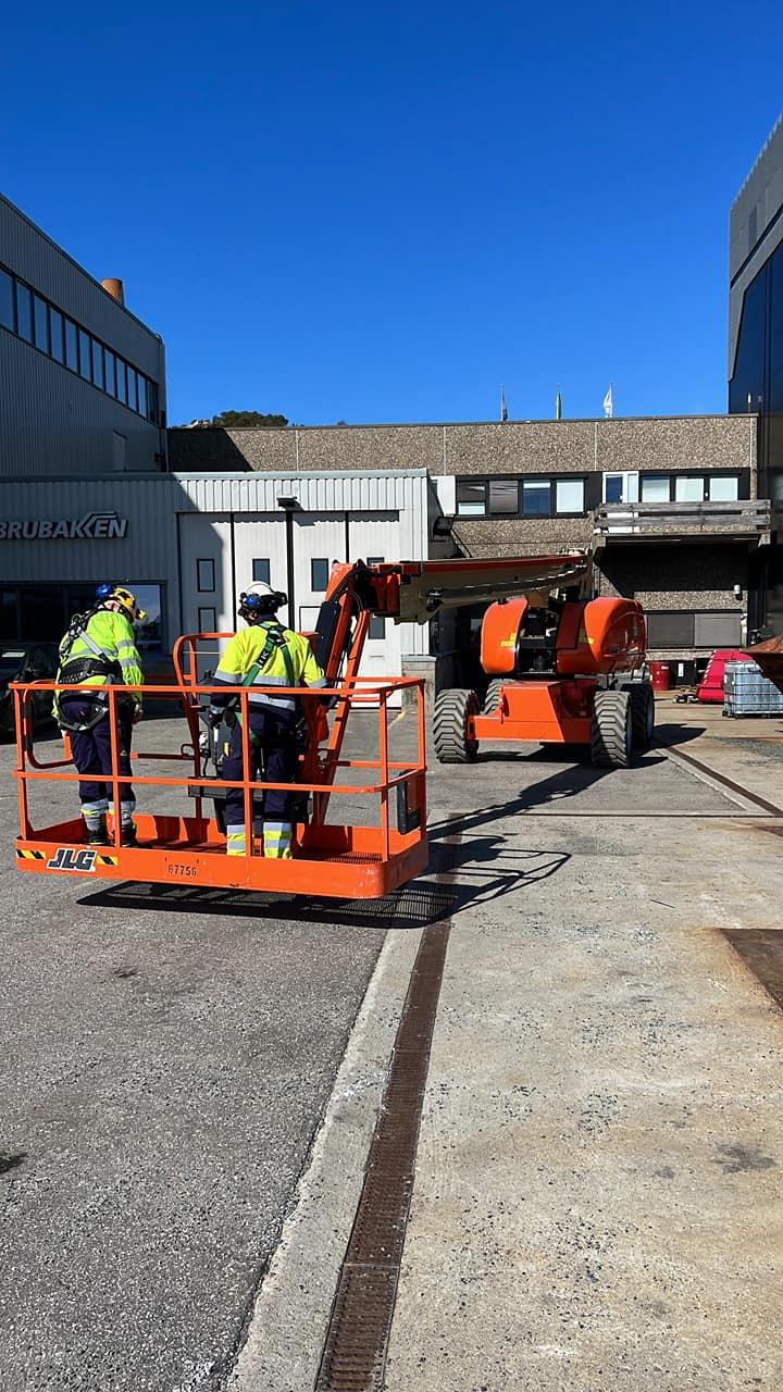 To personer i sikkerhetsutstyr står på en heis, mens en stor arbeidsmaskin er parkert i bakgrunnen.