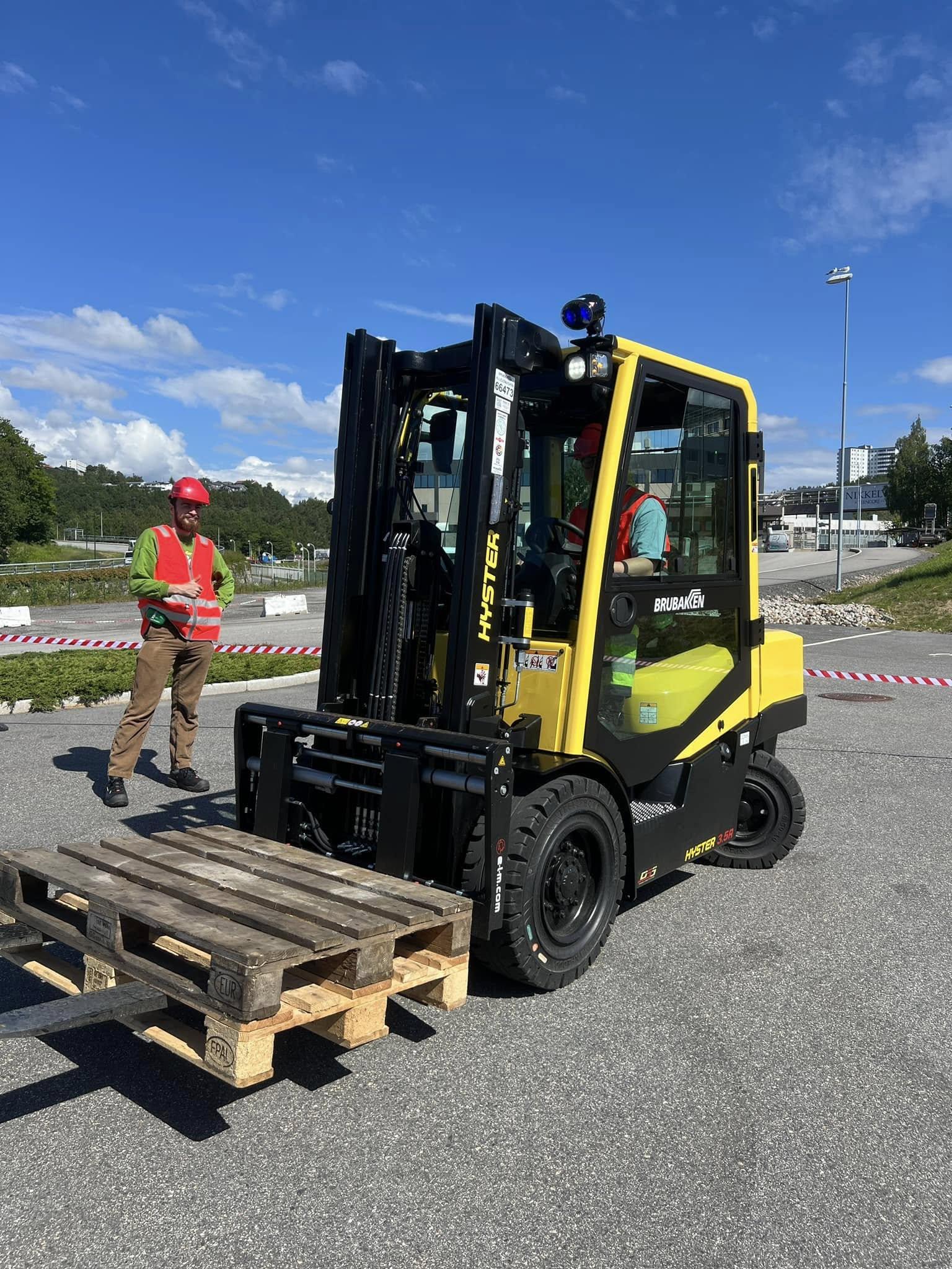Gul gaffeltruck løfter en pall på en byggeplass med en arbeider i rød hjelm i bakgrunnen.
