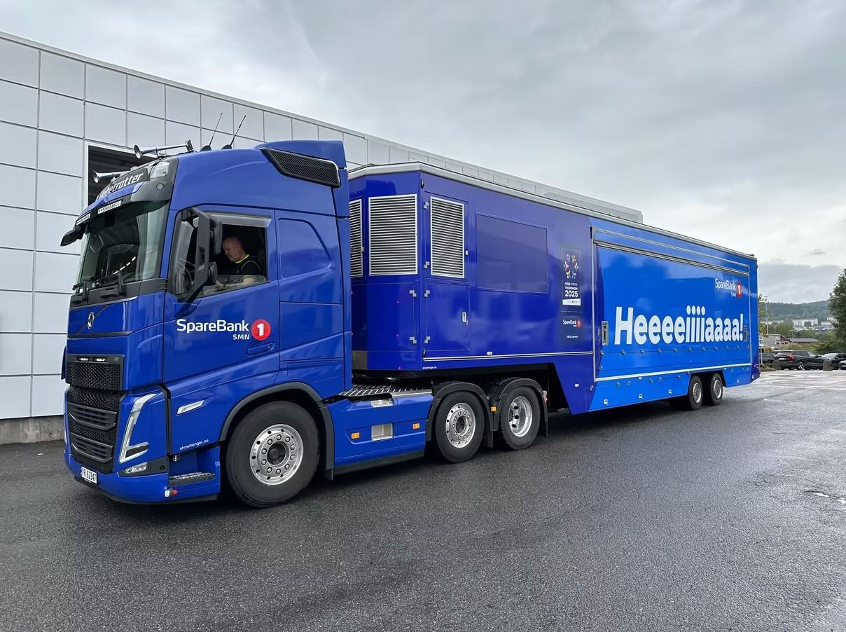 Blå lastebil med påhengsvogn, merket "Heeeelmaaaas", parkert foran en bygning.