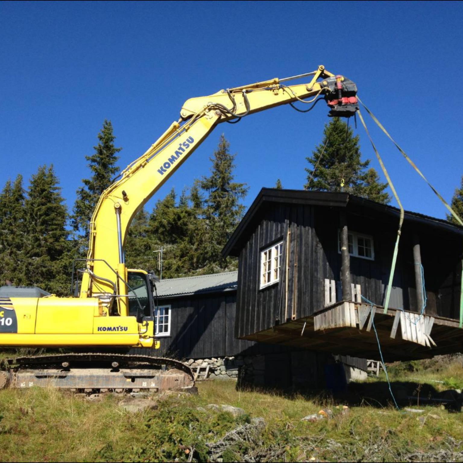 Gravemaskin løfter en hytte med kran i et skogsområde, omringet av trær og grønt gress.