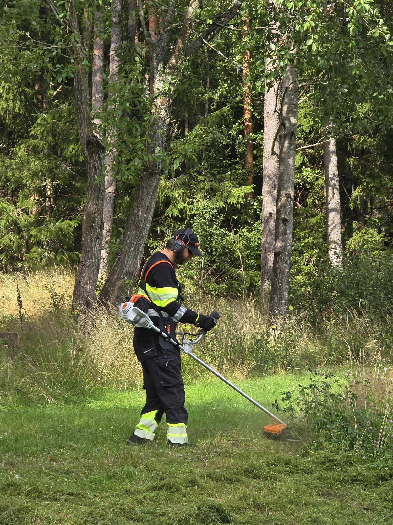 En mann i arbeidsklær bruker en gresstrimmer i en skogkledd hage med trær og høyt gress.