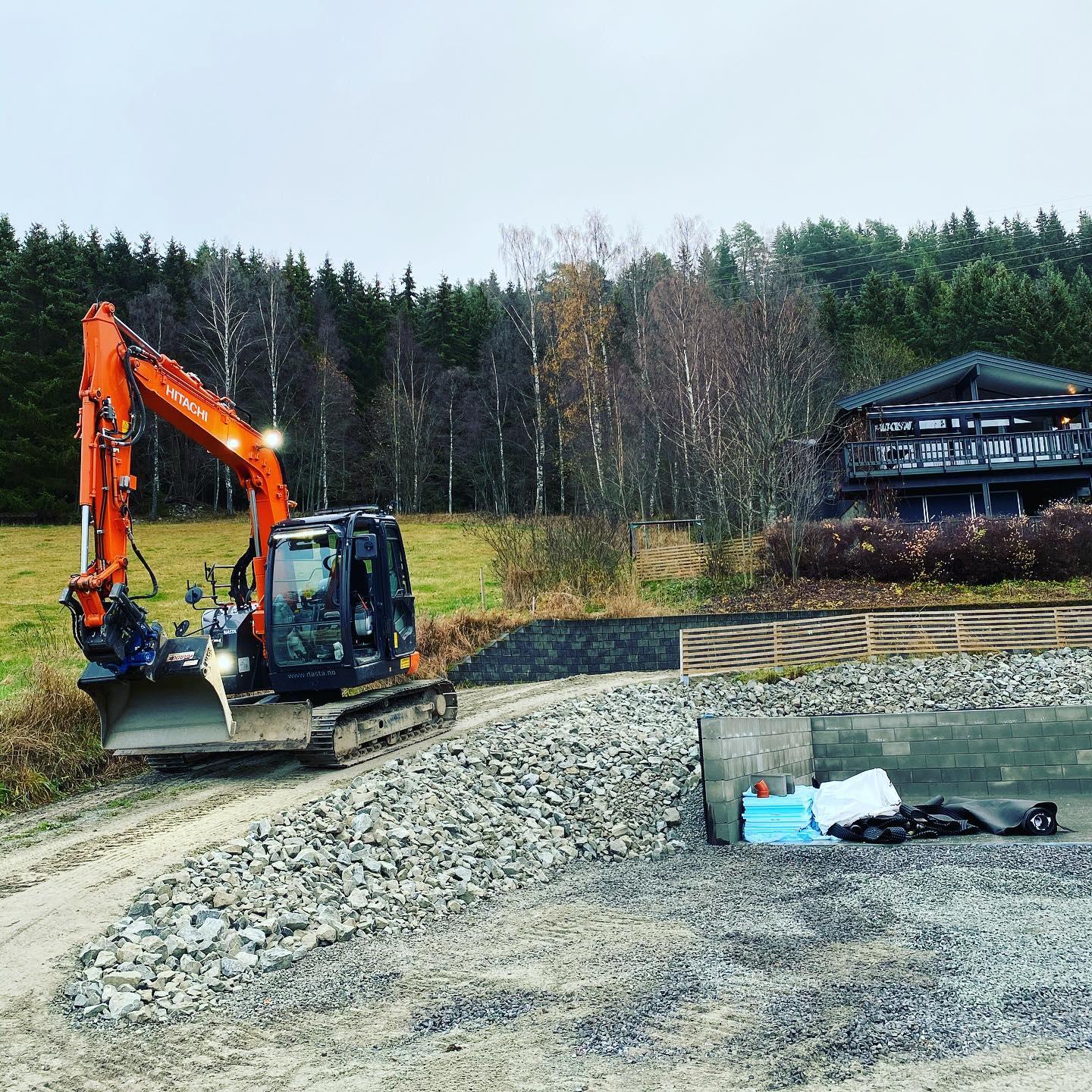 Gravemaskin arbeider på en byggeplass med en hytte i bakgrunnen og steinmateriale rundt.