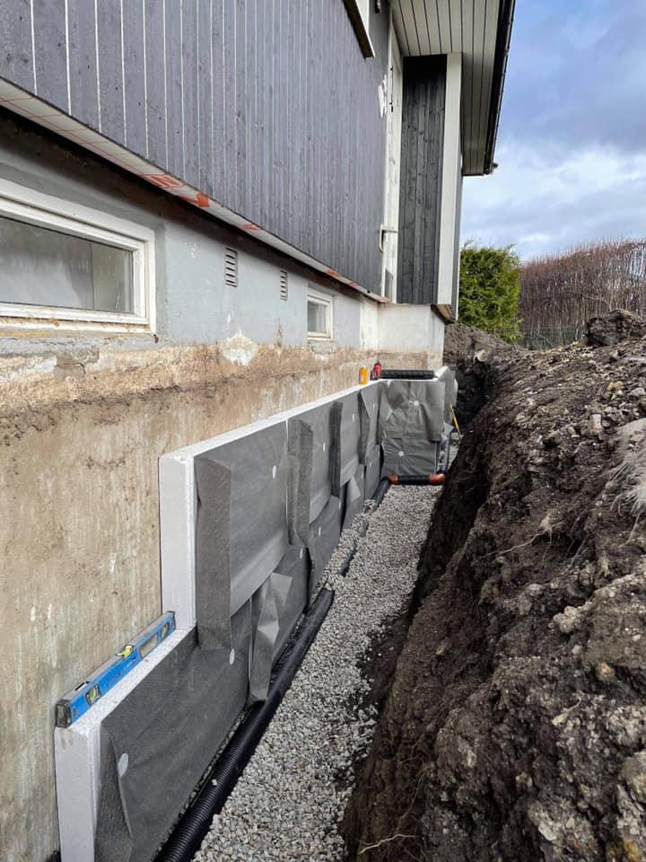 Grunnmur med drenering og grus under et hus, med synlig isolasjon og graving rundt.