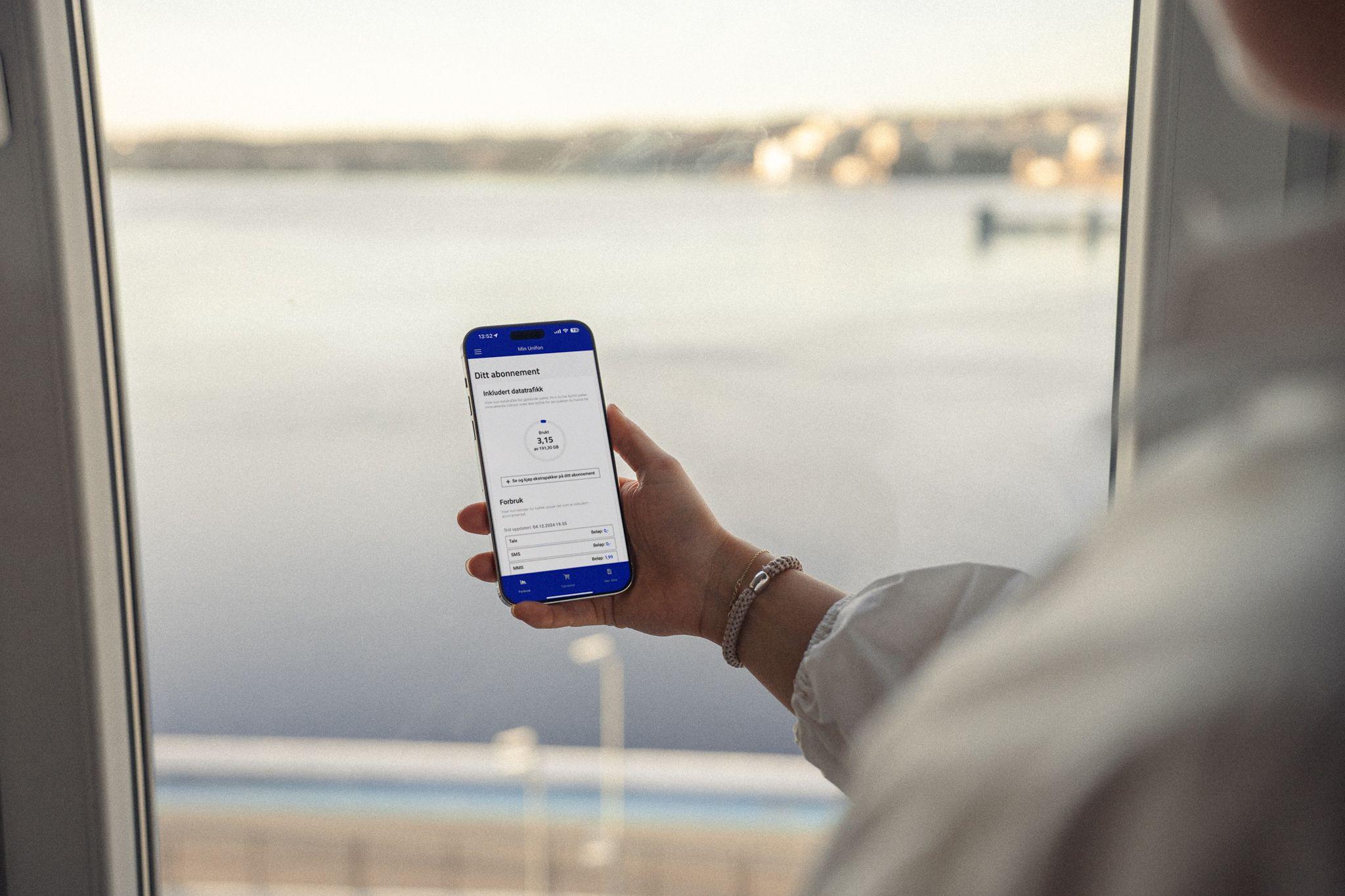 En person holder en smarttelefon med en app åpen, med utsikt over vannet i bakgrunnen.