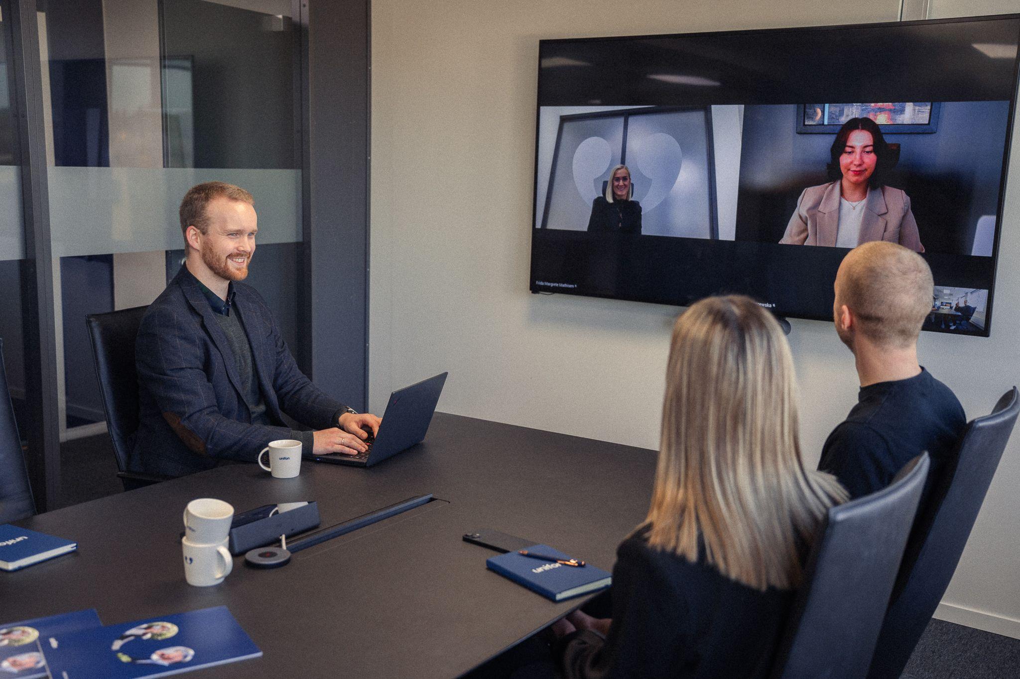 Møte i et konferanserom med fire personer, hvorav to deltar via videokonferanse på skjerm.