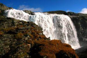 En vakker foss som renner nedover klippene, omringet av steiner og lyng under en klar blå himmel.