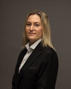 En kvinne med langt, lyst hår iført en svart dress og hvit skjorte, står med armene krysset.