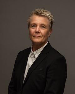 En person med kort, blondt hår iført en svart dress og hvit skjorte, står med armene lett krysset.
