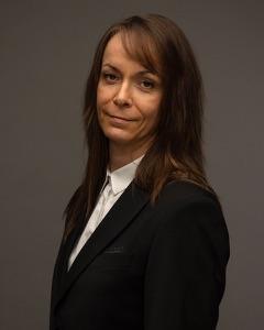 En kvinne med langt hår iført en svart dress og hvit skjorte, står med et seriøst uttrykk mot en grå bakgrunn.