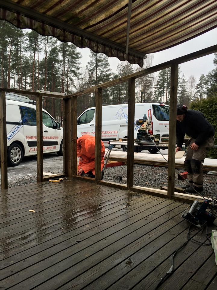 To personer jobber med å bygge en terrasse, med varebiler i bakgrunnen og trær rundt.