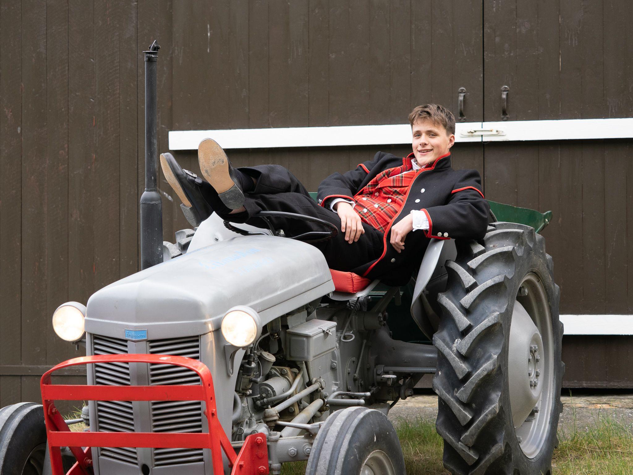 Ung mann i festdrakt som slapper av med bena på en grå traktor foran en brun vegg.