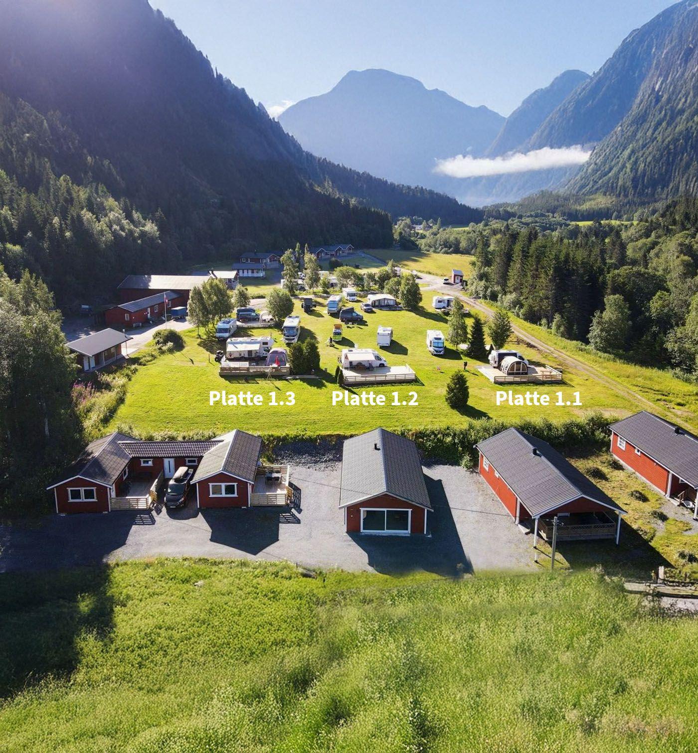 Oversiktsbilde av campingplass med hytter og telt i en dal, omringet av fjell og grønn natur.